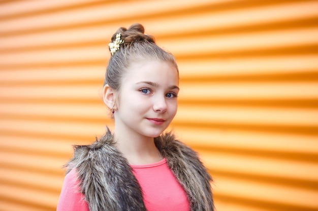 Portrait en gros plan d'une jolie petite fille souriante en robe rose et veste sans manches en fourrure posant près d'un mur rayé ondulé orange