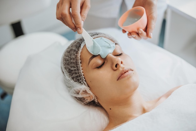 Portrait en gros plan des mains des cosmétologues appliquant un masque d'alginate avec une spatule sur le visage d'une femme en s...