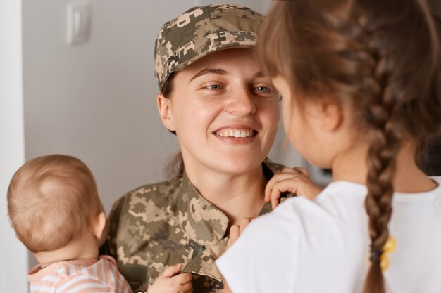 Portrait en gros plan d'une mère militaire souriante et optimiste portant un uniforme de camouflage et un chapeau, rencontrant ses filles lors du retour à la maison après le service militaire.