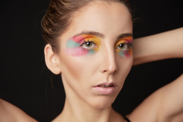 Portrait en gros plan d'un modèle blond avec un maquillage artistique très coloré. En studio avec un fond noir.