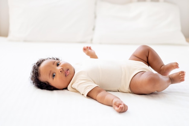 un portrait en gros plan d'une petite fille afro-américaine dans un body blanc sur un lit en coton à la maison un drôle de bébé nouveau-né noir souriant et joyeux de six mois se trouve sur le dos