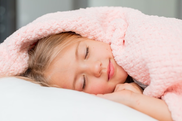 Portrait en gros plan d'une petite fille blonde allongée et dormant dans un lit sous une couverture rose