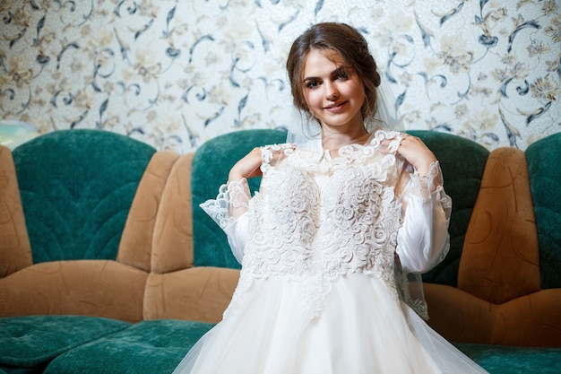 Portrait en gros plan photo d'une mariée avec un maquillage professionnel, elle est en blouse blanche, voile. Assis sur le canapé et regardant la robe de mariée. Jour de mariage de la jeune femme. Séance photo, coiffure, beauté