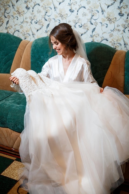 Portrait en gros plan photo d'une mariée avec un maquillage professionnel, elle est en blouse blanche, voile. Assis sur le canapé et regardant la robe de mariée. Jour de mariage de la jeune femme. Séance photo, coiffure, beauté