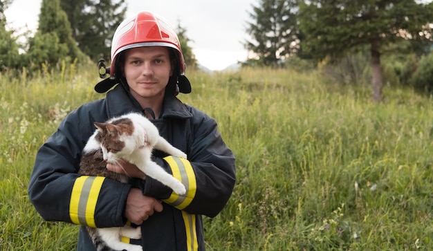 Portrait en gros plan d'un pompier héroïque en tenue de protection et casque rouge tenant un chat sauvé dans ses bras. Pompier en opération de lutte contre l'incendie. Photo de haute qualité