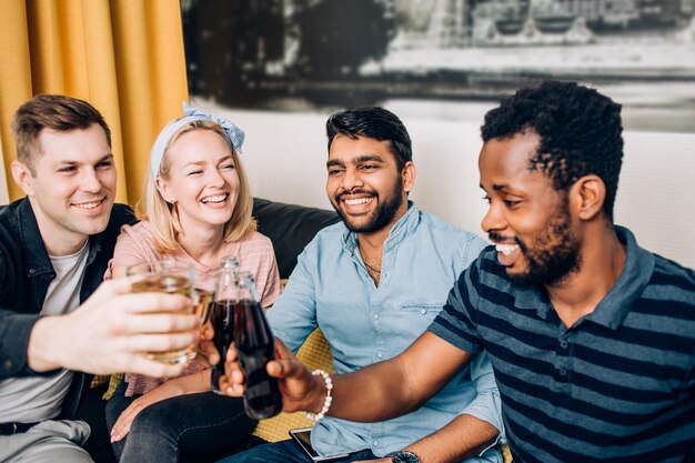 Portrait en gros plan de quatre amis joyeux faisant tinter leurs verres et bouteilles de boissons, souriant assis sur un canapé à la maison. Concept de fête à la maison, de célébration, de personnes et de vacances.