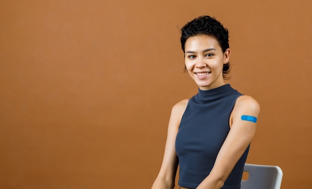 Portrait Gros plan en studio photo d'une jolie belle patiente sexy souriante montrant un bandage en plâtre bleu sur son épaule après avoir reçu la vaccination contre le coronavirus Covid-19 devant le mur marron.
