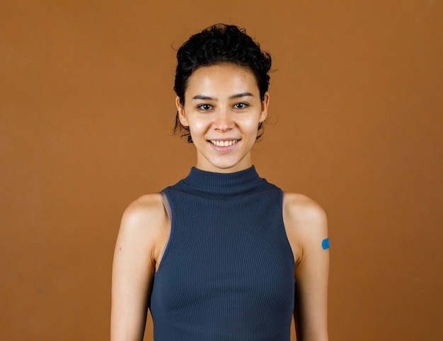 Portrait Gros plan en studio photo d'une jolie belle patiente sexy souriante montrant un bandage en plâtre bleu sur son épaule après avoir reçu la vaccination contre le coronavirus Covid-19 devant le mur marron.