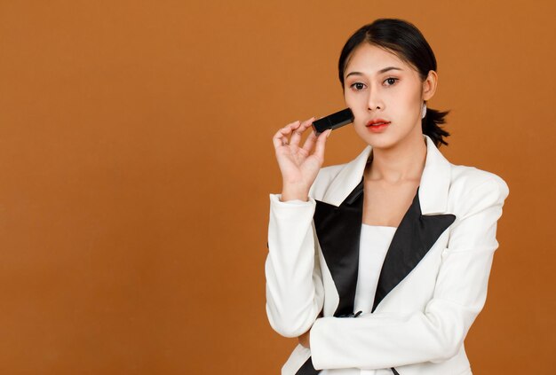 Portrait en gros plan tourné en studio Le modèle féminin asiatique de cheveux courts en queue de cheval noire du millénaire porte du maquillage en blazer décontracté de mode noir et blanc, bras croisés tenant du rouge à lèvres rouge à la main sur fond marron.