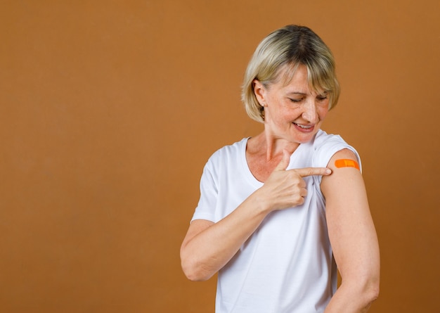 Portrait Gros plan tourné en studio d'une patiente blonde âgée de race blanche, soulignant qu'elle souffre d'une blessure par injection au pansement de plâtre orange sur son épaule après avoir reçu la vaccination Covid-19.