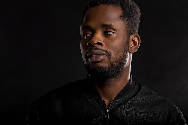 Portrait en gros plan très détaillé d'un jeune homme africain séduisant portant une veste noire regardant la caméra avec une expression sérieuse et réfléchie sur fond sombre. Émotions et sentiments humains.