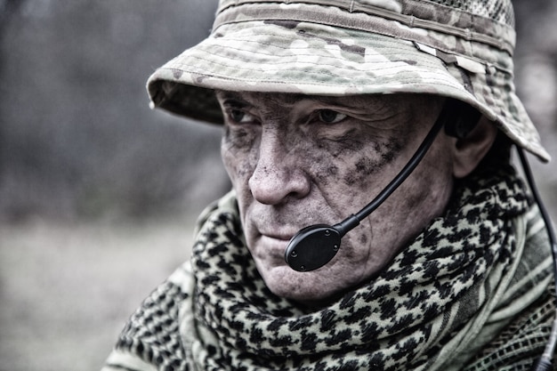 Photo portrait en gros plan d'un vétéran du commando brutal, d'un commandant de l'armée expérimenté ou d'un officier au visage sale, portant une bonnie de camouflage, un shemagh, un casque radio tactique avec microphone, regardant à huis clos