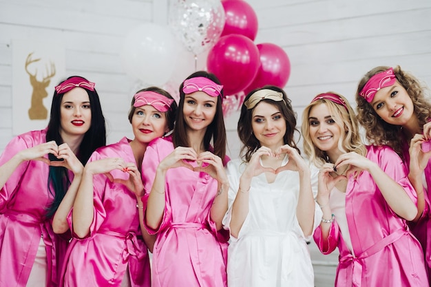Portrait d'un groupe de copines heureuses en robes rose vif et masques de sommeil montrant des cœurs avec les mains lors de la célébration de la douche nuptiale