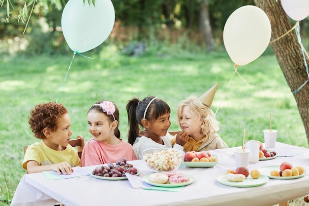 Portrait d'un groupe diversifié d'enfants à table de pique-nique à l'extérieur profitant d'une fête d'anniversaire en été