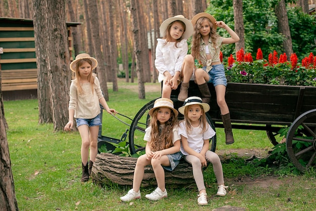 Portrait de groupe de préadolescentes dans des vêtements d'été décontractés et des chapeaux de soleil en osier marchant dans un parc de la ville verte posant près d'un vieux chariot en bois rustique utilisé comme lit de fleurs avec de la salvia rouge en fleurs