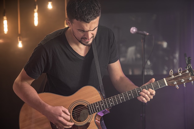 Portrait de guitariste souriant en t-shirt noir jouant de la guitare acoustique sur un espace sombre studio flou.