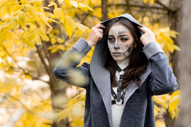 Portrait d'Halloween Une belle jeune fille vêtue d'une robe blanche avec du maquillage sur son visage dans un manteau à capuchon se tient effrayant dans la forêt d'automne Le jour des morts