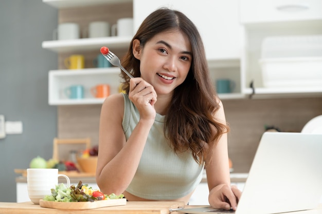 Portrait Happy Asia woman look at camera avec salade et ordinateur portable dans la salle de cuisine