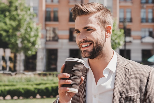 Portrait de l'heure du café d'un homme aux cheveux bruns confiant tenant une tasse de café et souriant de côté en marchant