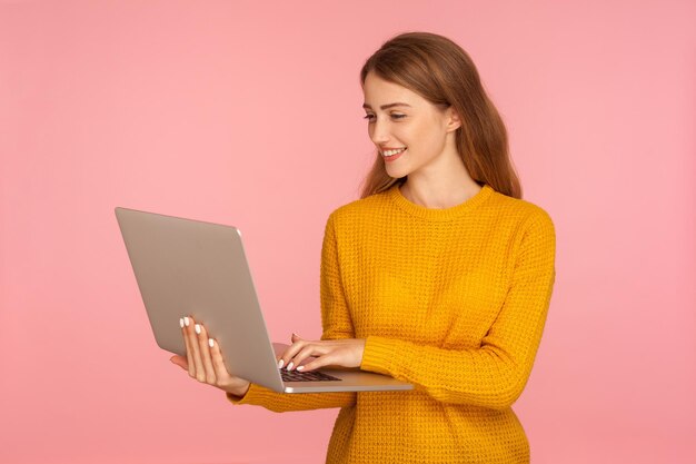 Portrait d'une heureuse fille au gingembre positive en chandail tenant un ordinateur portable, tapant sur le clavier, surfant sur Internet, étudiant ou pigiste travaillant à l'aide d'un ordinateur. studio d'intérieur tourné isolé sur fond rose