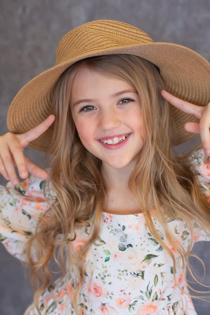 portrait d'une heureuse fille blonde aux cheveux longs dans une robe à fleurs et un chapeau de paille sur fond gris