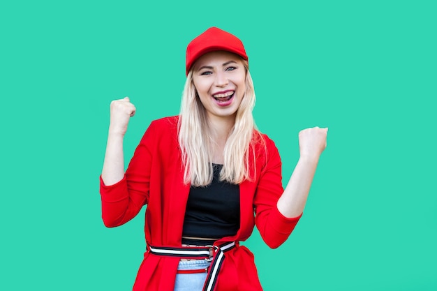 Portrait de l'heureuse gagnante belle femme blonde de style hipster jeune en blouse rouge et casquette, debout regardant la caméra et célébrant sa victoire. tourné en studio intérieur, isolé sur fond vert.