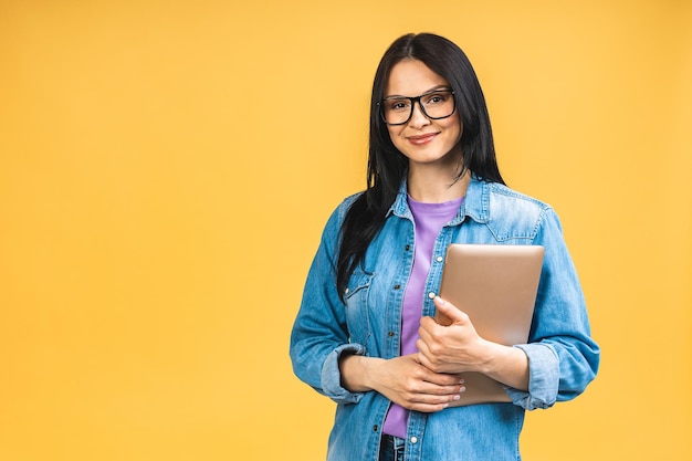 Portrait d'heureuse jeune belle femme surprise avec des lunettes debout avec un ordinateur portable isolé sur fond jaune Espace pour le texte