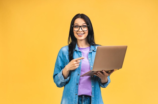 Portrait d'heureuse jeune belle femme surprise avec des lunettes debout avec un ordinateur portable isolé sur fond jaune Espace pour le texte