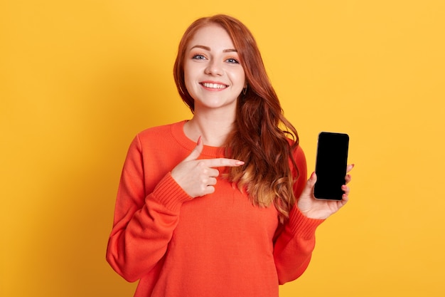 Portrait de l'heureuse jeune femme de race blanche pointant sur un écran vide de téléphone portable dans sa main isolé sur espace jaune