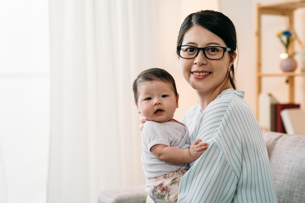 portrait heureuse mère de carrière asiatique tenant un mignon bébé innocent regarde la caméra avec un sourire dans un salon confortable avec lumière du jour.