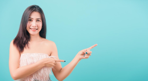 Portrait heureux asiatique belle jeune femme debout son doigt pointé à l'extérieur et regardant la caméra isolée sur fond bleu avec espace de copie pour le texte