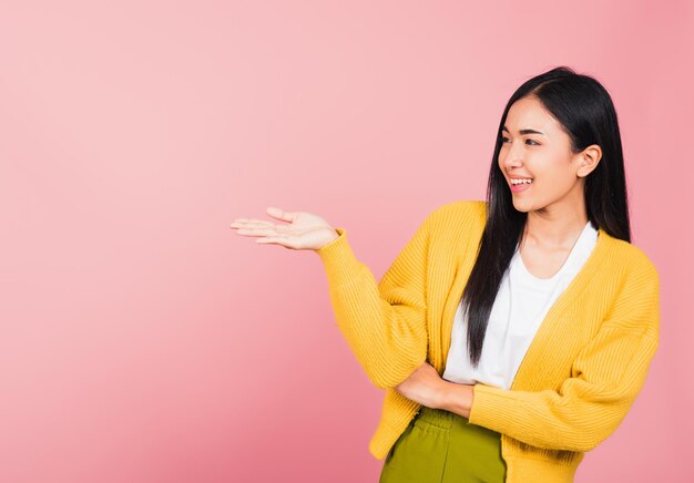 Portrait heureux asiatique belle jeune femme mignonne sourire debout présentant le produit tenant quelque chose sur le côté de la paume, prise de vue en studio isolée sur fond rose avec espace de copie, geste de la main du spectacle féminin