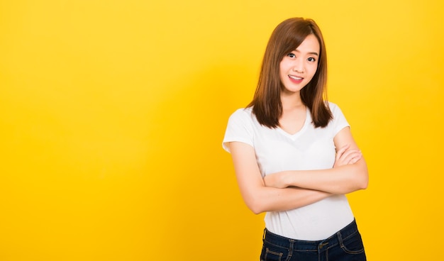 Portrait heureux asiatique belle jolie jeune femme adolescente debout porter un t-shirt sourire sa confiance avec les bras croisés à la recherche d'appareil photo isolé, studio tourné sur fond jaune avec espace de copie de bannière