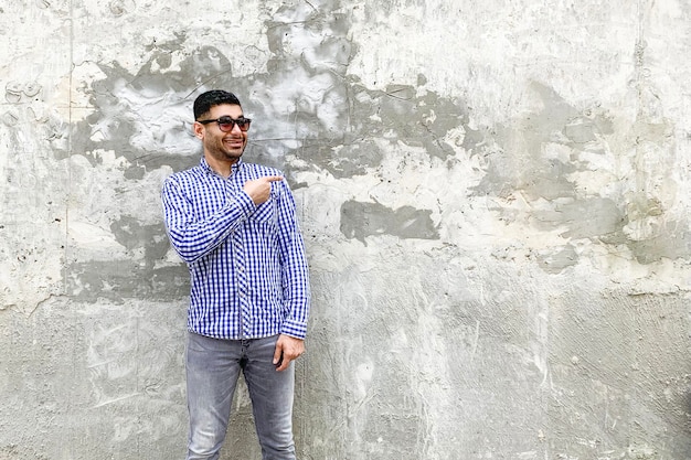 Portrait d'heureux beau jeune homme barbu en chemise bleue à carreaux et lunettes de soleil debout contre un mur gris en béton pointant vers un fond vide et regardant la caméra avec un sourire à pleines dents