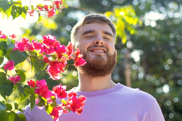 Portrait d'heureux beau mec barbu jeune homme positif avec barbe sent de belles fleurs roses rouges dans le jardin souriant appréciant le printemps ou l'été respirant profondément l'air frais