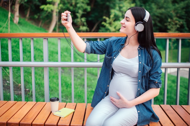 Portrait d'un heureux cheveux noirs et fière femme enceinte dans le parc. Elle est assise sur un banc de ville. La future femme écoute de la musique dans le parc avec un enfant à naître