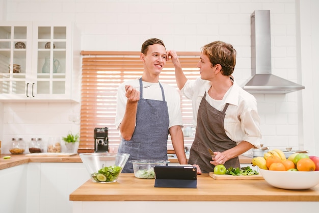 Portrait de l'heureux couple gay caucasien portant des tabliers se regardant avec des sourires et appréciant de cuisiner ensemble dans la cuisine à la maison Relations LGBT Concept de cuisine de couple gay