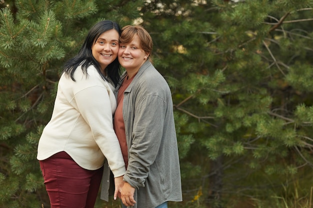 Portrait de l'heureux couple de lesbiennes embrassant et souriant à la caméra en se tenant debout dans la forêt en journée d'été