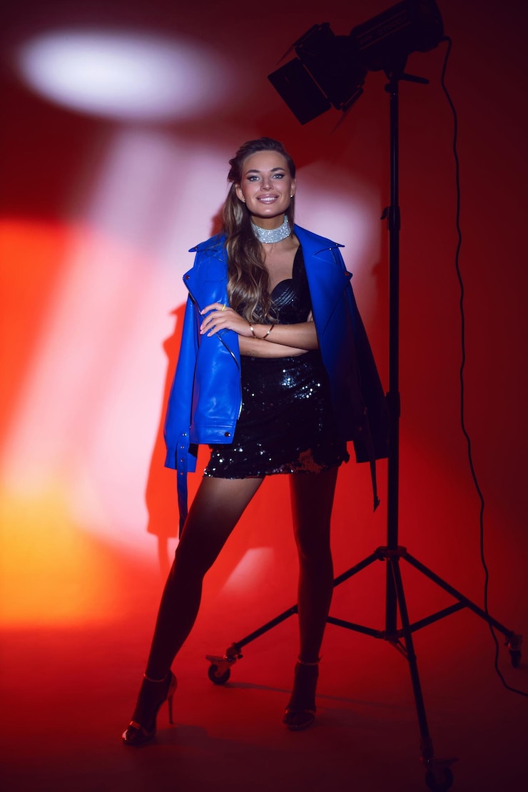 Photo portrait heureux d'une femme sexy dans une robe bottes et veste bleue se tient dans le studio sur un cyclorama sur un fond rouge avec un dispositif de lumière