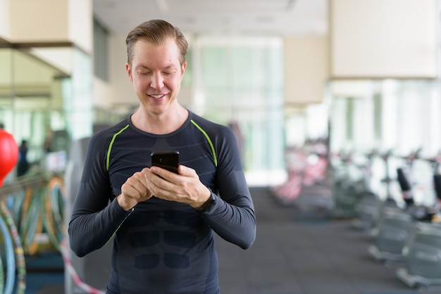 Portrait d'heureux jeune bel homme à l'aide de téléphone à la salle de sport pendant covid-19