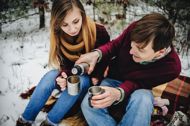 Portrait de l'heureux jeune couple au pique-nique le jour de la Saint-Valentin dans un parc enneigé. L'homme et la fille se versent dans des tasses de vin chaud, du thé chaud, du café avec thermos. Vacances de Noël, fête.