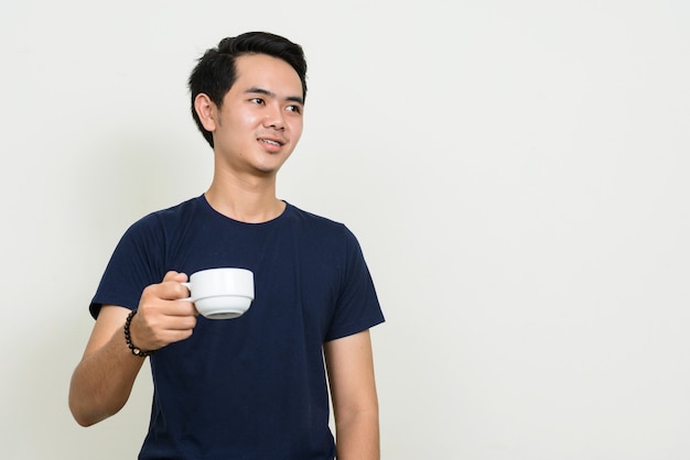 Portrait d'heureux jeune homme asiatique pensant tout en tenant une tasse de café