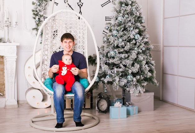 Portrait d'heureux jeune père assis dans une chaise et jouant avec son bébé près de l'arbre de Noël.