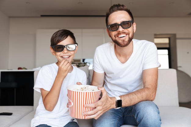 Portrait de l'heureux père et fils portant des lunettes 3D, manger du pop-corn et souriant, assis sur un canapé à l'intérieur et regarder un film