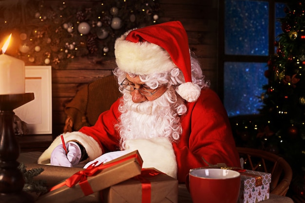 Portrait de l'heureux Père Noël assis dans sa chambre à la maison près de l'arbre de Noël et répondant aux lettres de Noël.