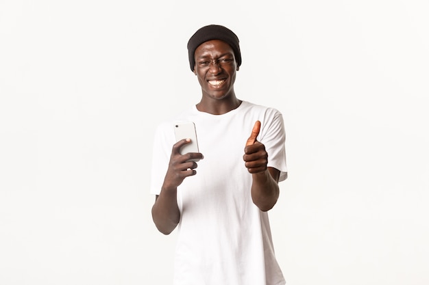 Portrait d'heureux et satisfait, beau mec afro-américain souriant largement, montrant les pouces vers le haut après avoir utilisé l'application de téléphone mobile