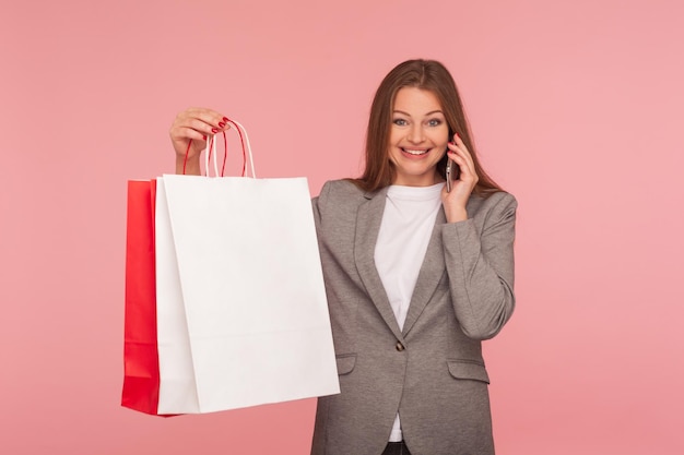 Portrait, de, heureux, shopper, client affaires, élégant, femme, dans, veste costume, tenue, sacs provisions, et, parler téléphone, sourire, semble, satisfait, à, achat, projectile studio, isolé, sur, rose, fond