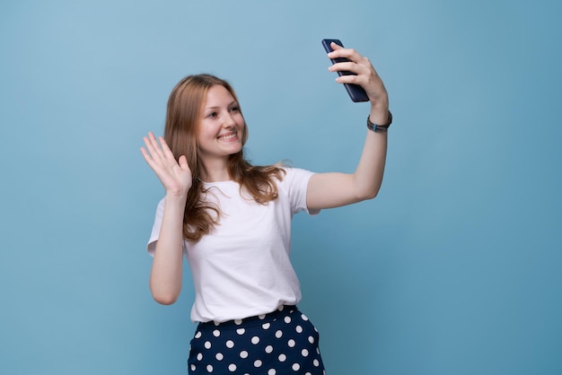 Portrait heureux sympathique jeune fille caucasienne en t-shirt blanc sur fond bleu prenant selfie et wa...