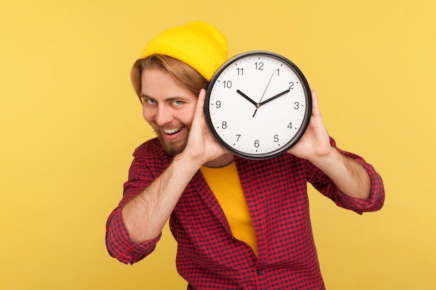 Portrait d'un hipster joyeux et positif en chemise à carreaux tenant une grande horloge et regardant la caméra avec un sourire à pleines dents, une expression optimiste ludique. studio d'intérieur tourné isolé sur fond jaune