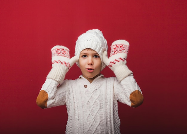 Portrait d'hiver d'un enfant joyeux et joyeux garçon dans un bonnet tricoté et un pull s'amusant sur fond rouge le garçon regarde ses paumes